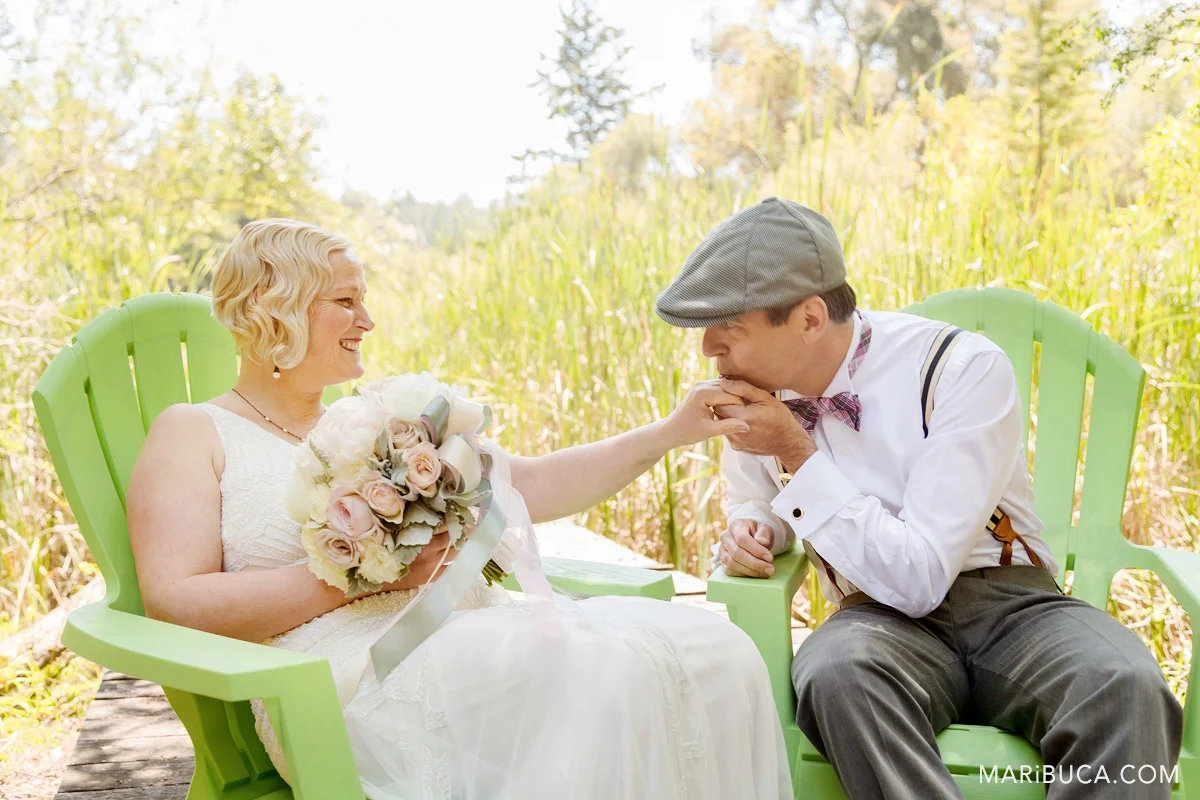 The groom kisses the bride's hand after the ceremony and they sit in the light green comfy chairs and high grass surrounded.