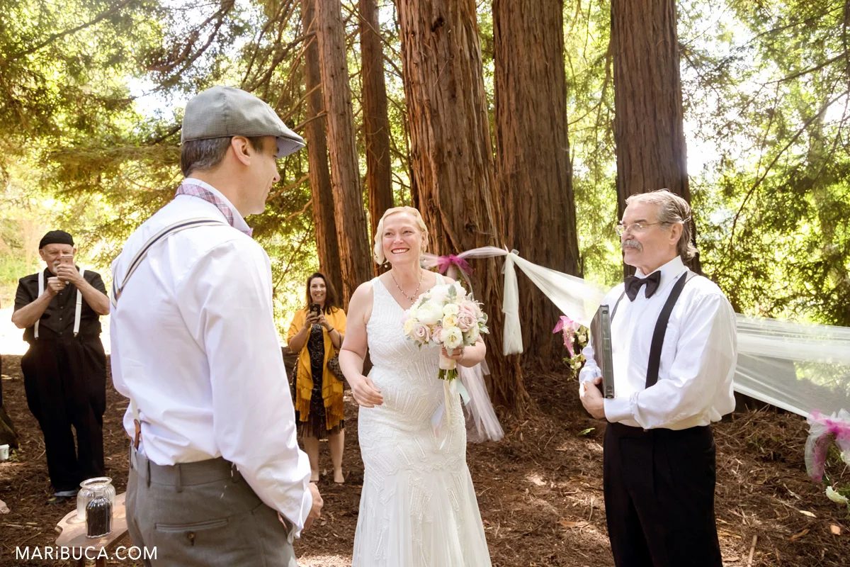 The bride is looking at the groom and smiling during wedding ceremony in the wood.
