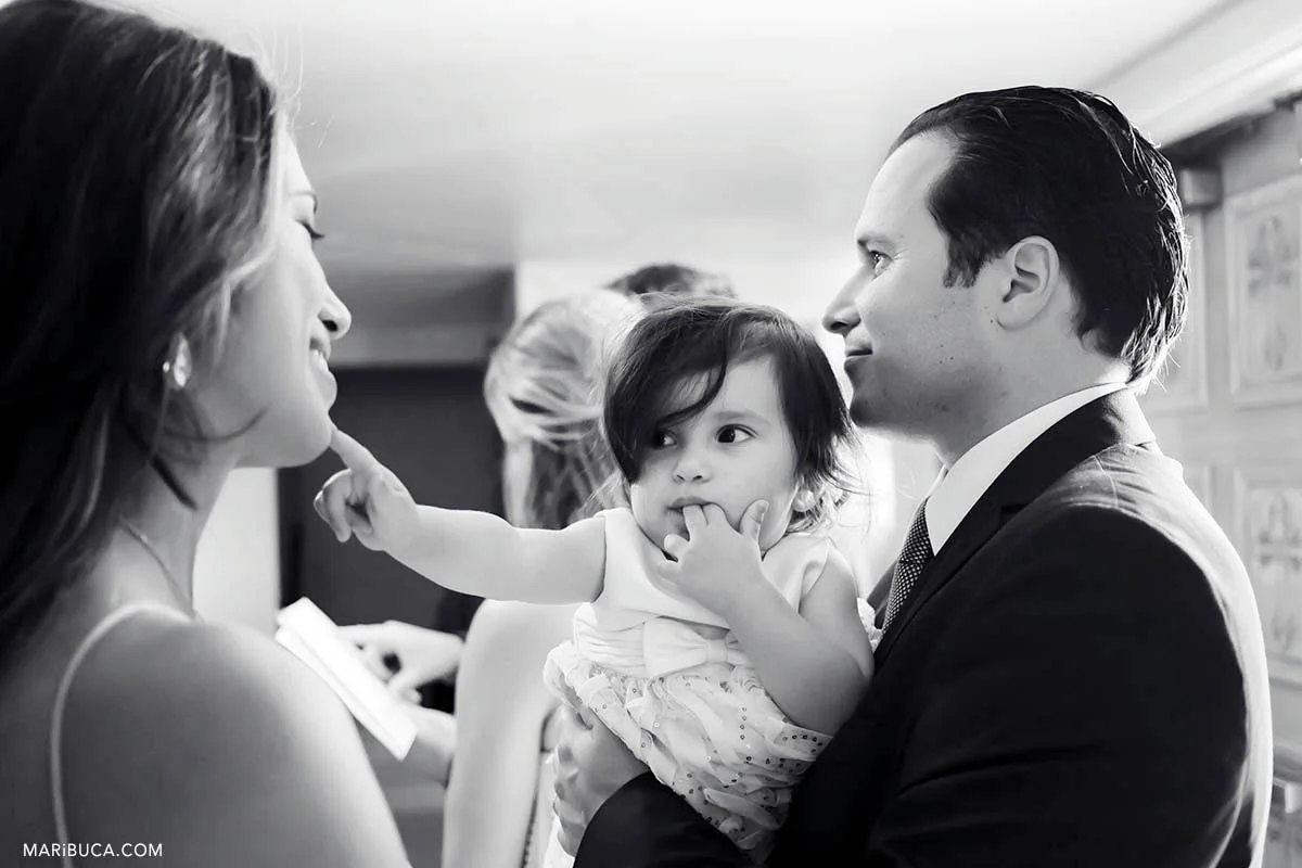 Black and white image. The baby girl is touching mom and mother smiling. Dad is listening the priest during baptism ceremony.