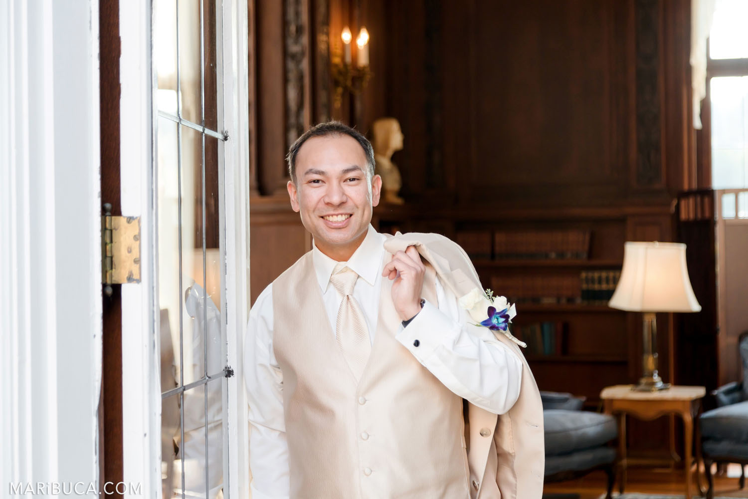 The groom holds wedding light beige jacket through the shoulder and stays in the Library, Kohl Mansion, Burlingame