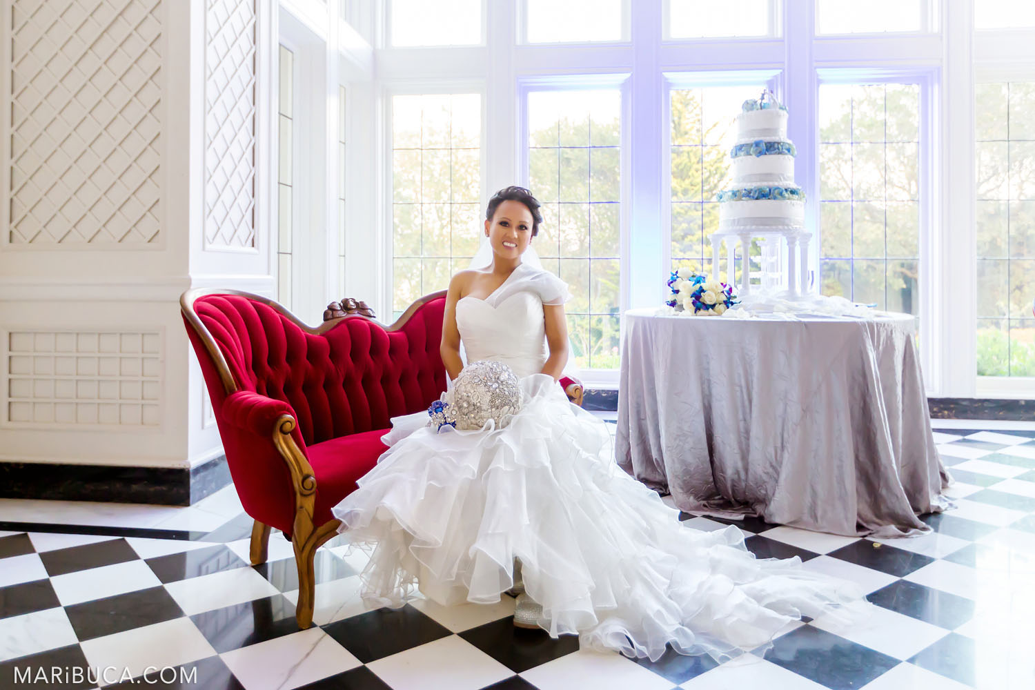 Gorgeous bride sits in the red couch around the Morning Room with big high windows, white walls and white three-stores wedding cake in the Kohl Mansion, Burlingame