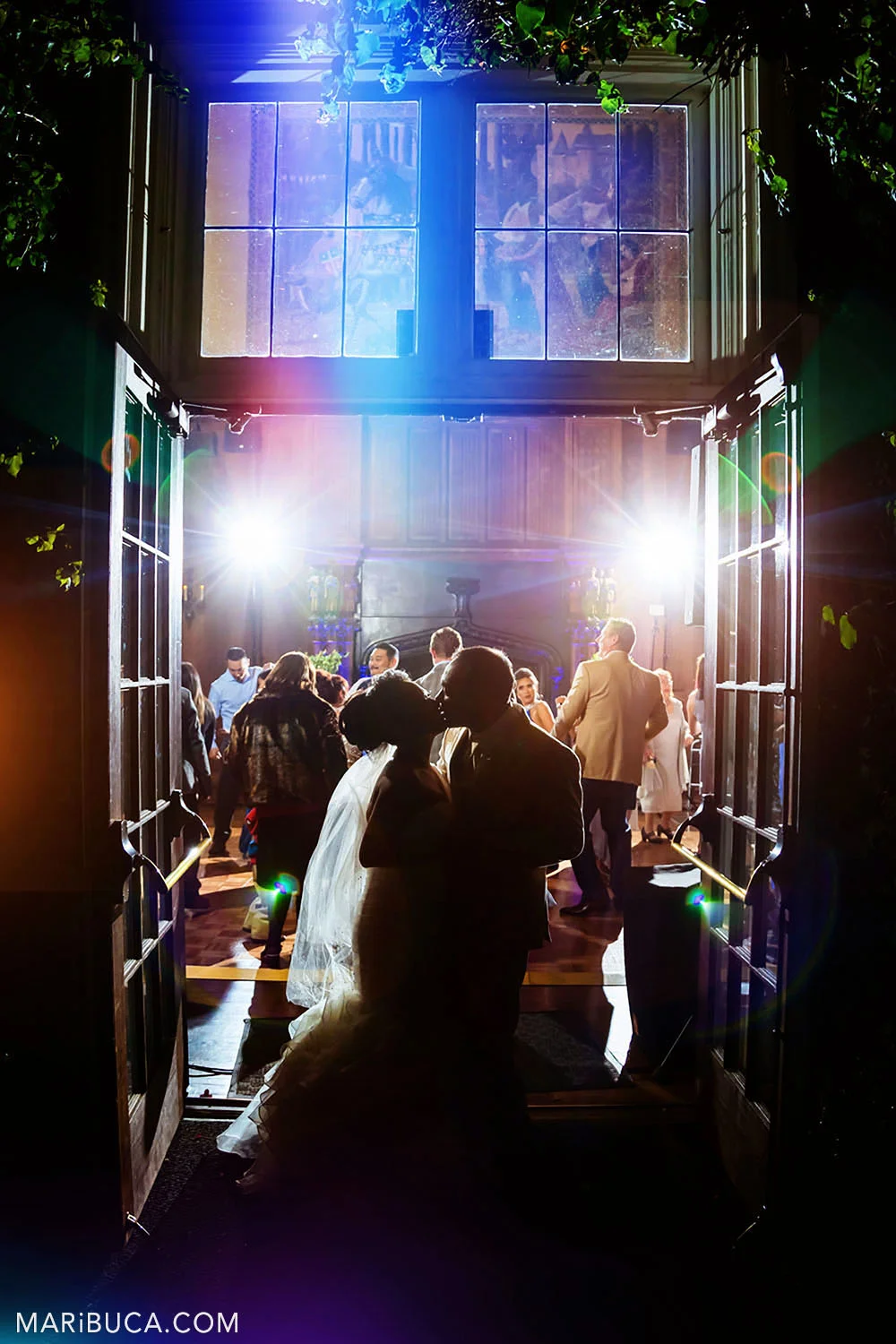 Bride and groom kiss each other surrounded by colorful lights and guests in the Great Hall, Kohl Mansion, Burlingame