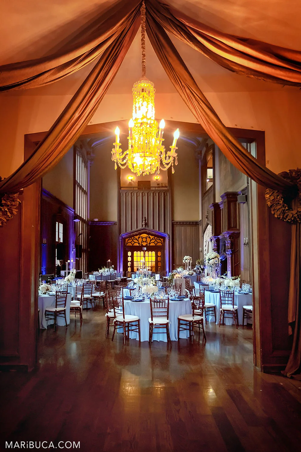 Wedding decoration of the Great Hall with brown walls and high ceilings such as round tables and light blue tablecloths, white fresh flowers on them and a purple backlight in the background in the Kohl Mansion, Burlingame