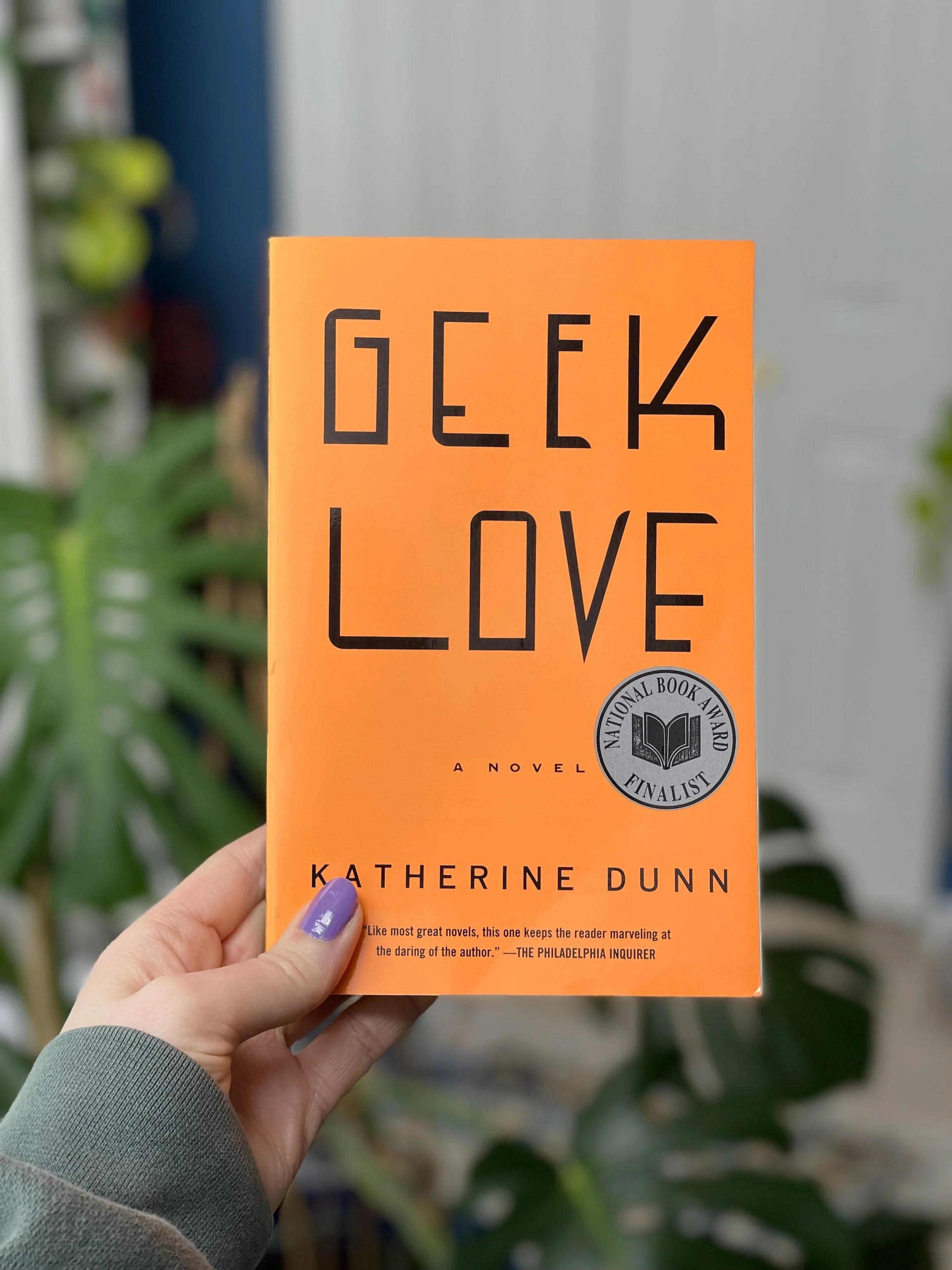 My hand holds a paperback copy of Geek Love by Katherine Dunn. The cover is a bright orange with black text.