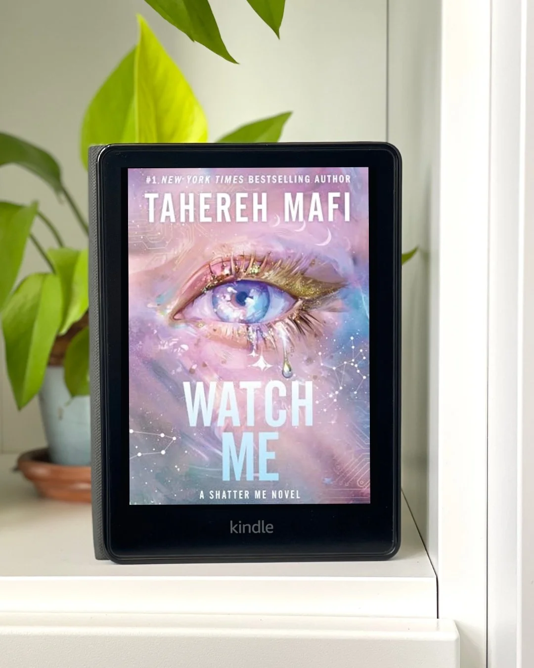 My Kindle sits on a white bookshelf with the cover of Watch Me by Tahereh Mafi on the screen. The cover is a light pink with a purple eye in the center.