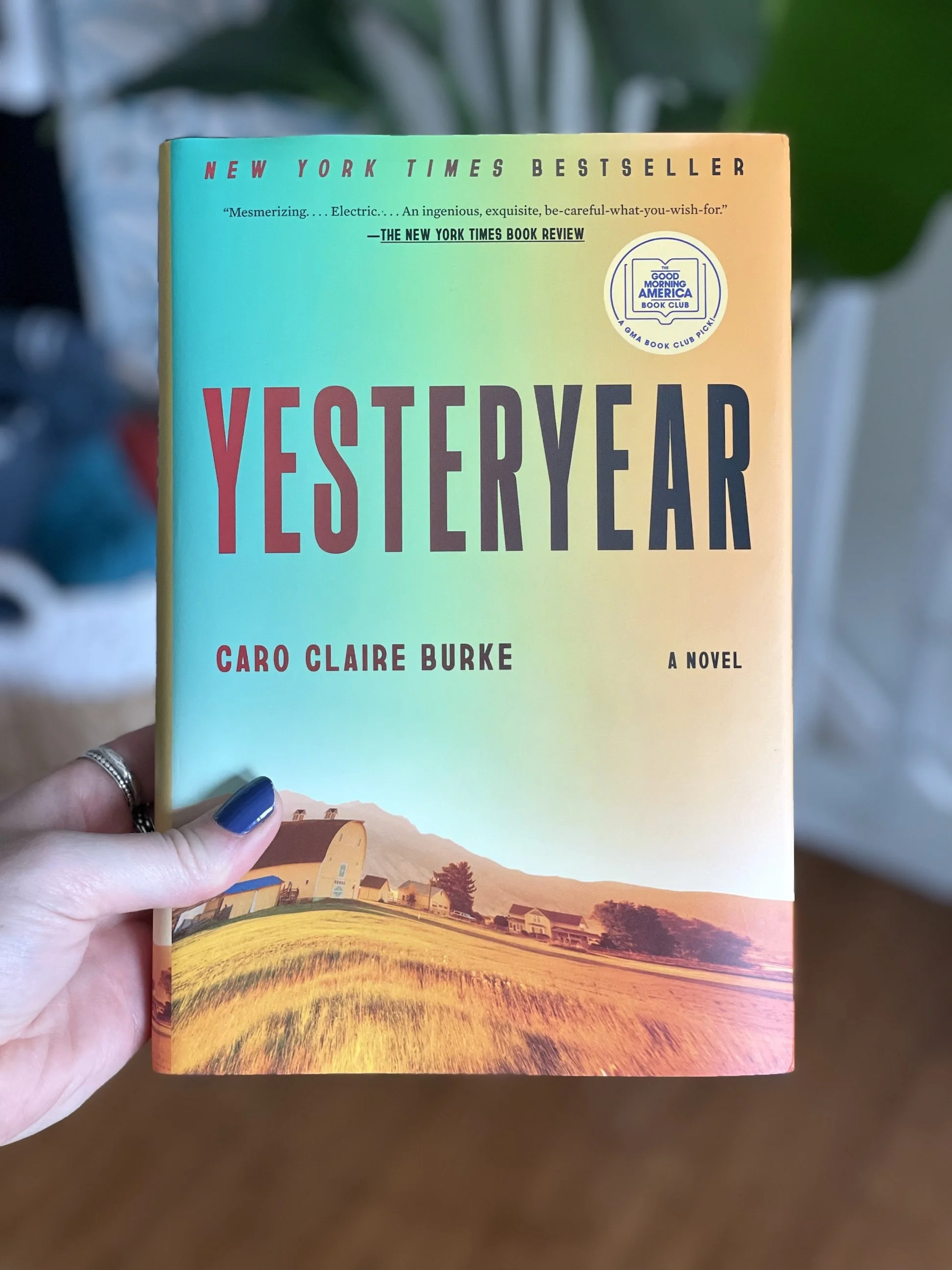 My hand holds a hard cover copy of Yesteryear by Caro Claire Burke. The cover is yellow and blue and depicts a small farm.