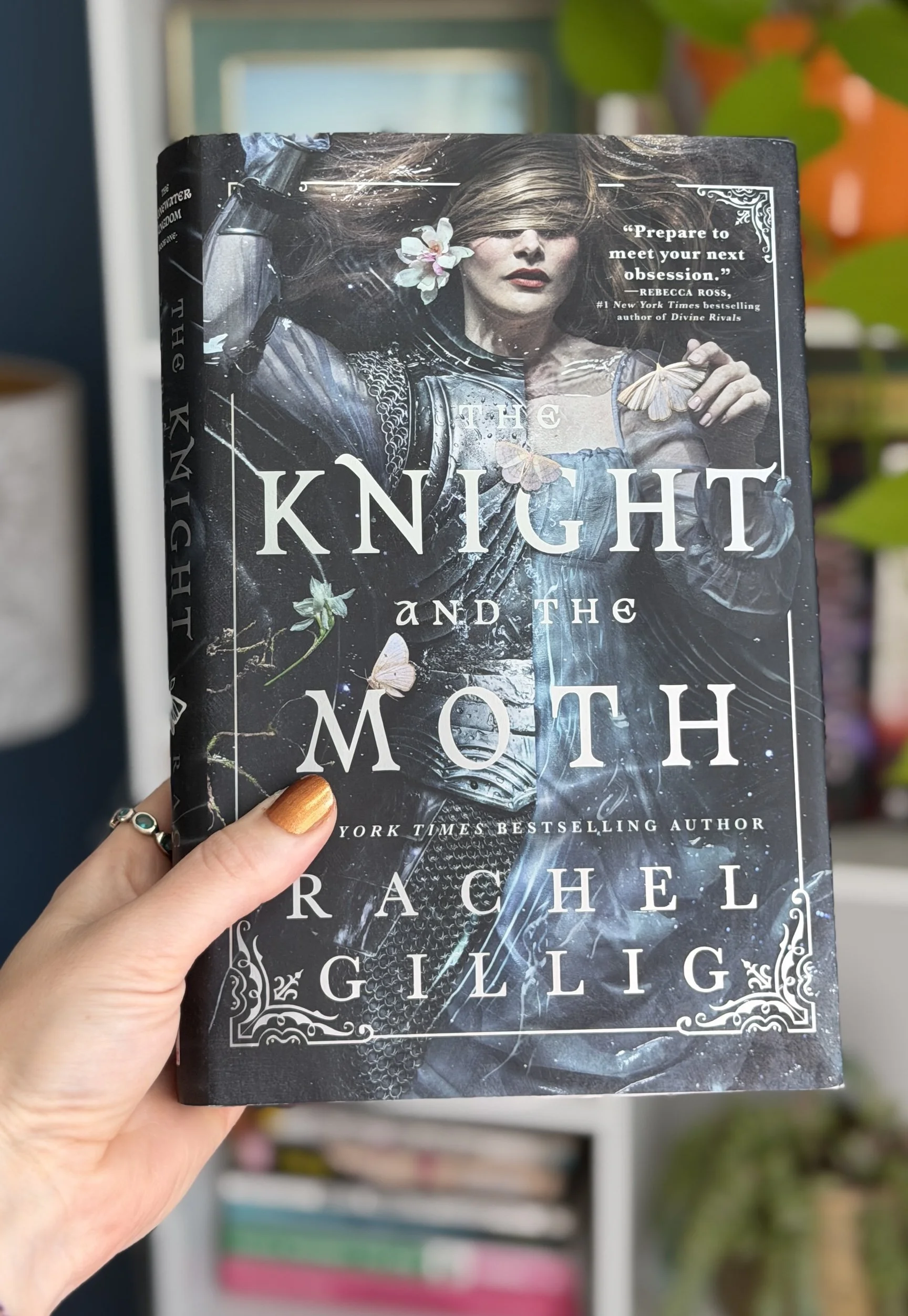 My hand holds a copy of The Knight and the Moth by Rachel Gillig. THe cover shows a woman in armor floating with a shroud over her eyes.
