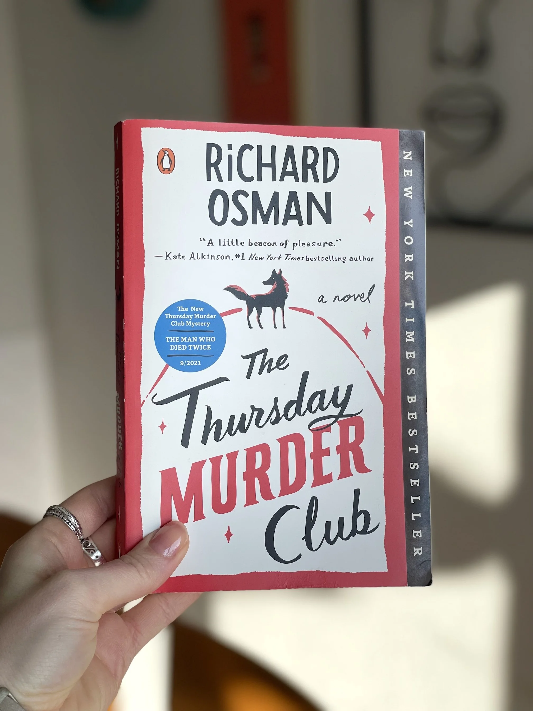 My hand holds a paperback copy of The Thursday Murder Club by Richard Osman. The cover is mostly red, white, and black, and has a dog on it.