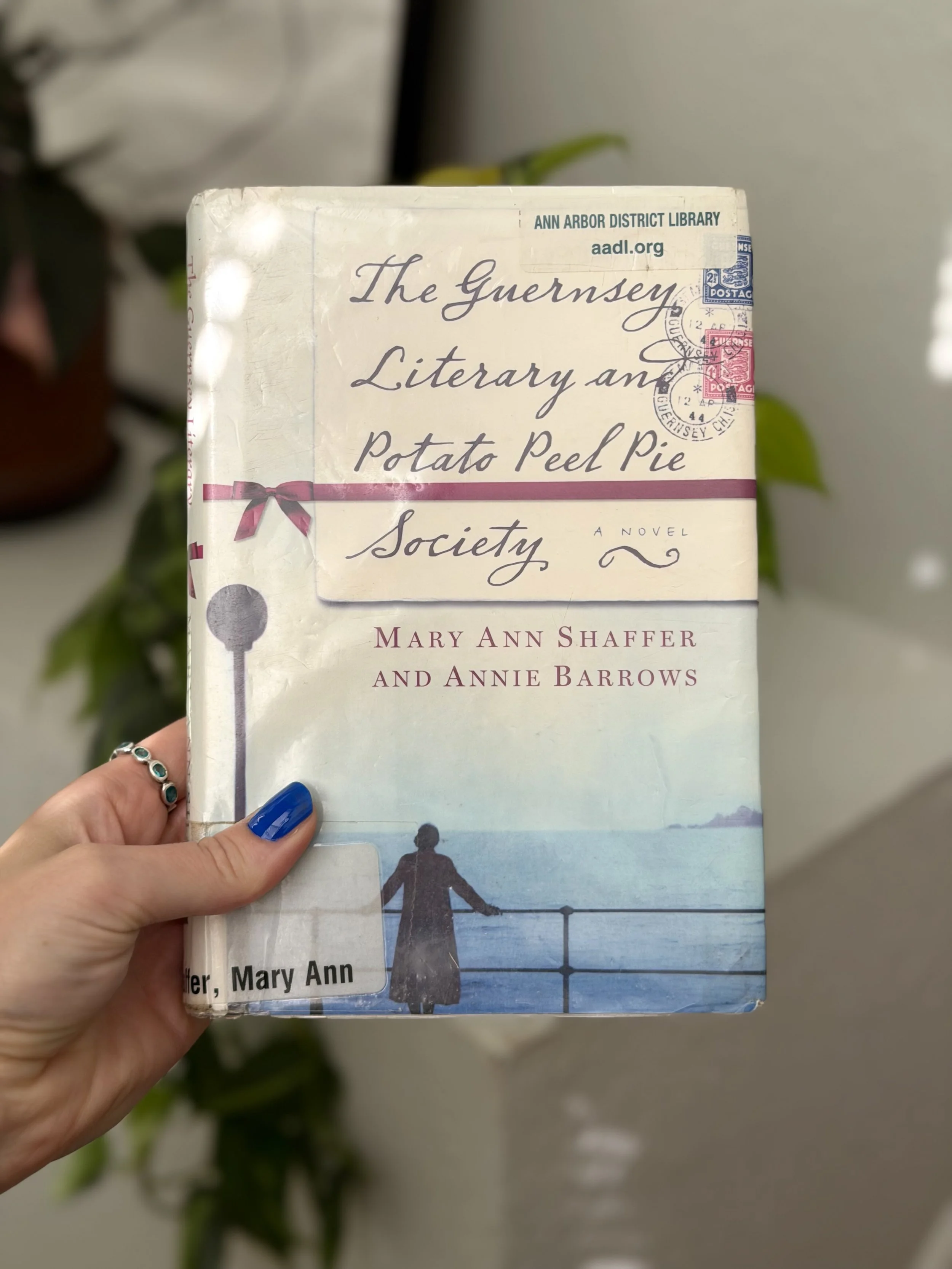 My hand holds a hard cover copy of The Guernsey Literary and Potato Peel Pie Society by Mary Ann Shaffer.