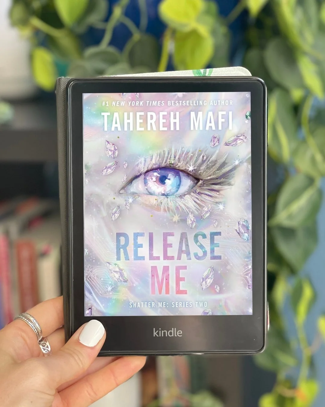 My hand holds a Kindle with Release Me by Tahereh Mafi on the screen. The cover is mostly silvery rainbow with a purple eye in the middle.