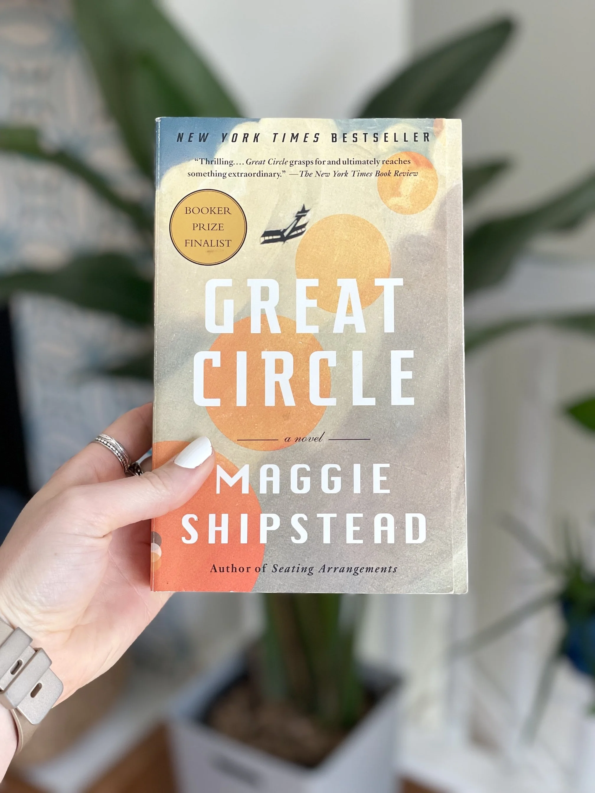My hand holds a paperback copy of Maggie Shipstead. The cover shows a sky with a plane flying through it.