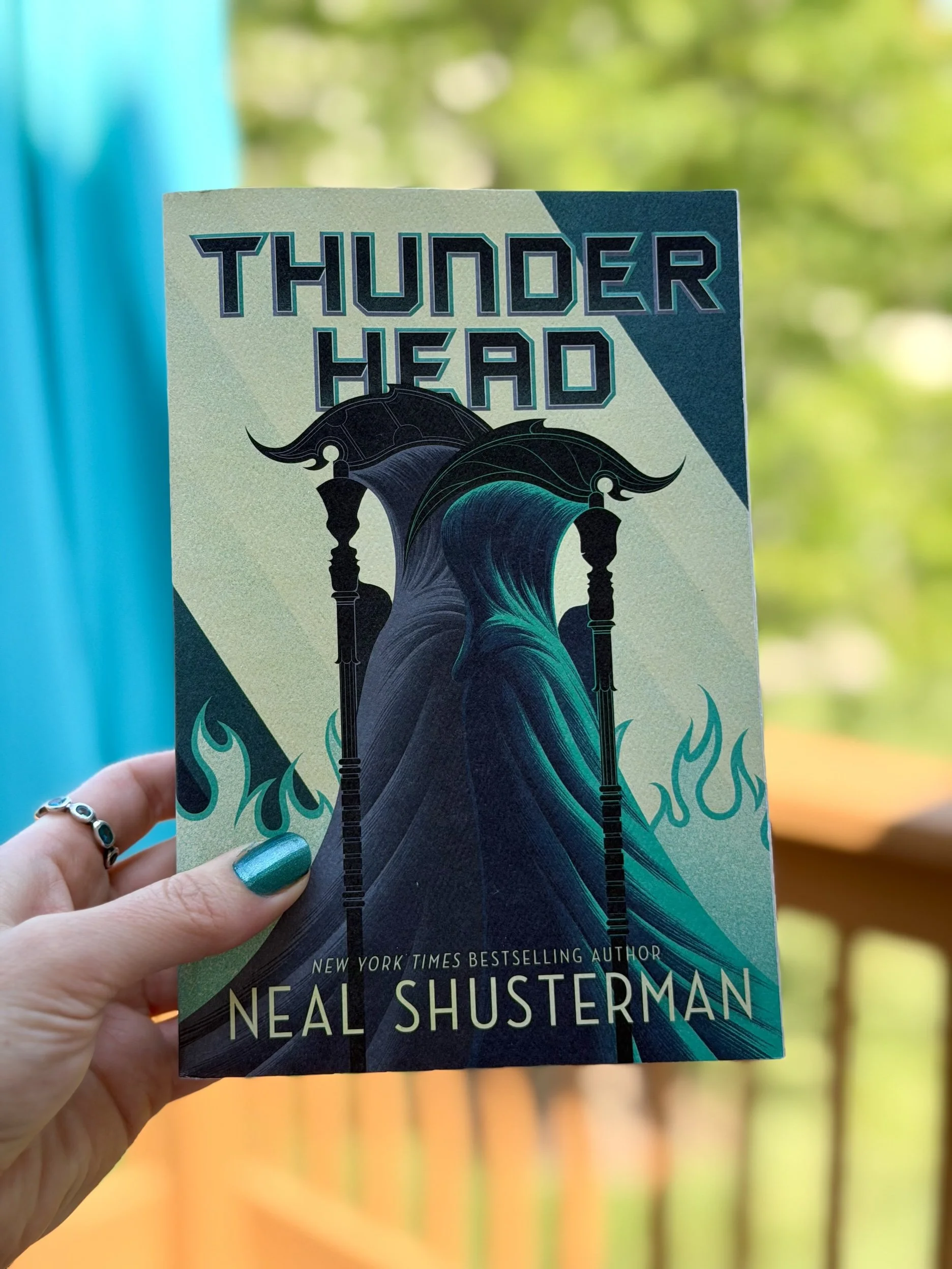 My hand holds a copy of Thunderhead by Neal Shusterman. The cover is mostly green with three figures in the middle.