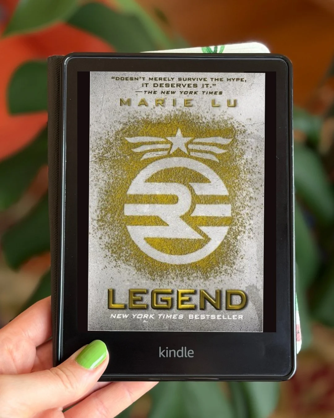My hand holds a kindle with the cover of Legend by Marie Lu on the screen. The cover is mostly white and gold with a symbol in the middle.