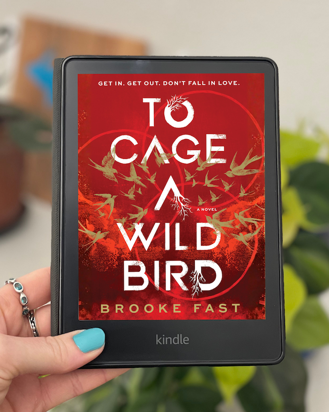 My hand holds a Kindle with the cover of To Cage a Wild Bird by Brooke Fast on the screen.