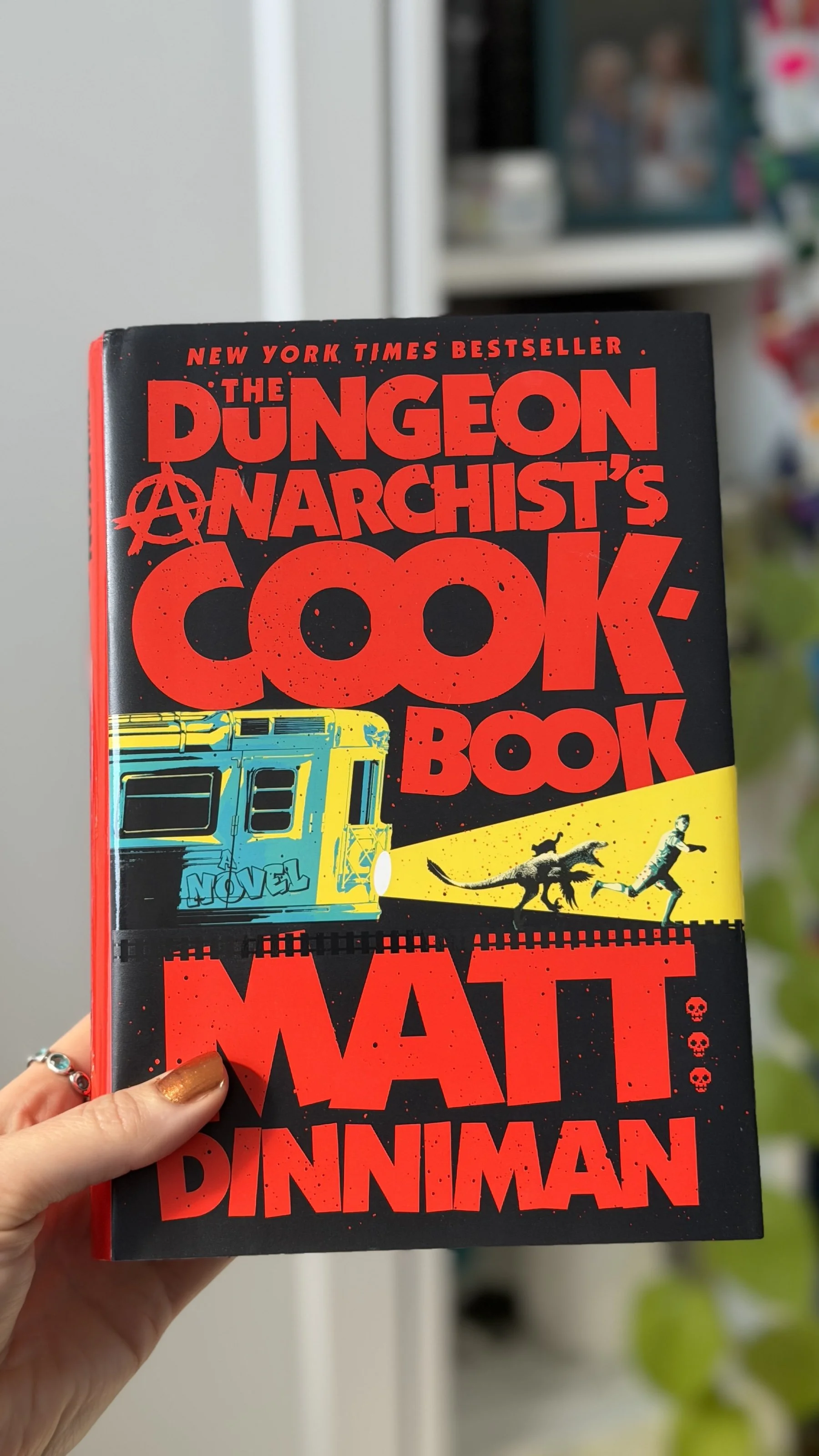 My hand holds a hardcover copy of The Dungeon Anarchist's Cookbook by Matt Dinniman which has a train car chasing a dinosaur, cat, and man on it.