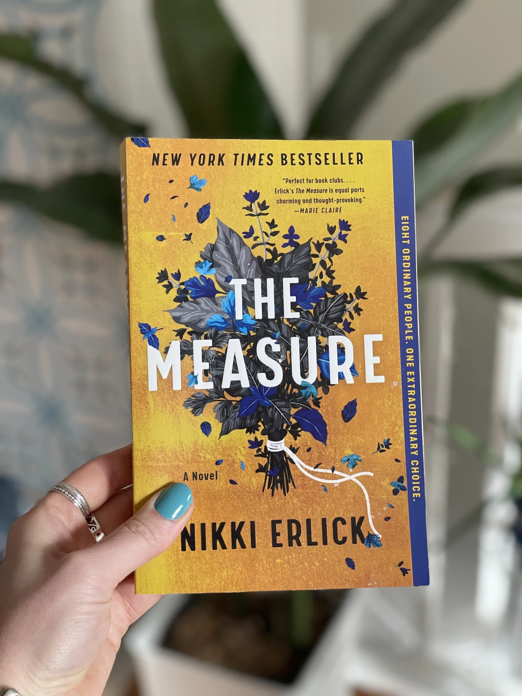 My hand holds a paperback copy of The Measure by Nikki Erlick. The cover is mostly yellow with a bouquet of blue and gray leaves in the center.