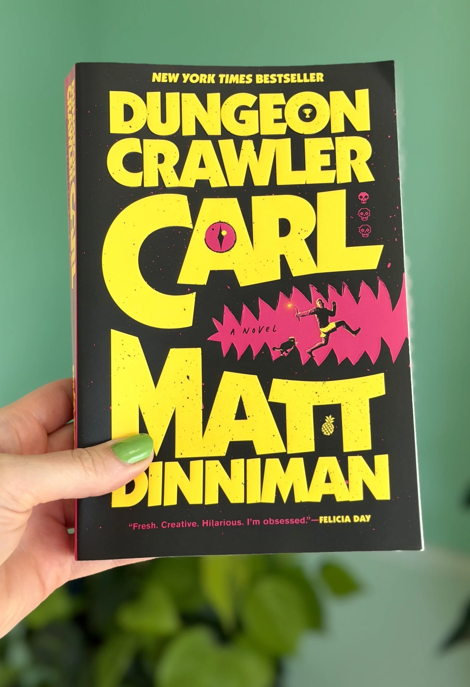 My hand holds a paperback copy of Dungeon Crawler Carl by Matt Dinniman. It's mostly black with yellow text and pink details.