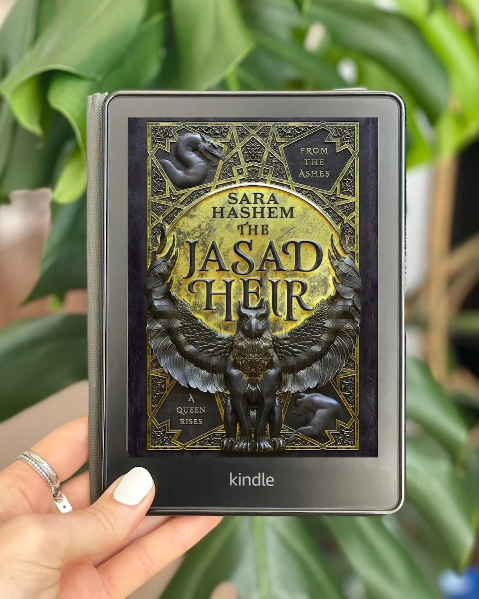 My hand holds a Kindle with The Jasad Heir by Sara Hashem on the screen. The cover shows a griffon statue in the center.