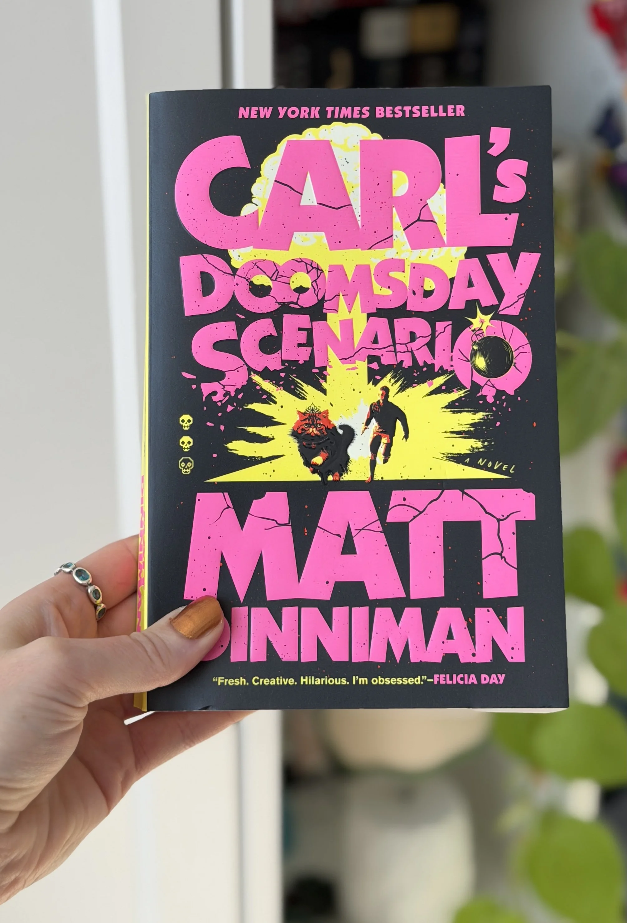 My hand holds a paperback copy of Carl's Doomsday Scenario by Matt Dinniman. The cover is black, yellow, and pink with a man and cat in the center of it.