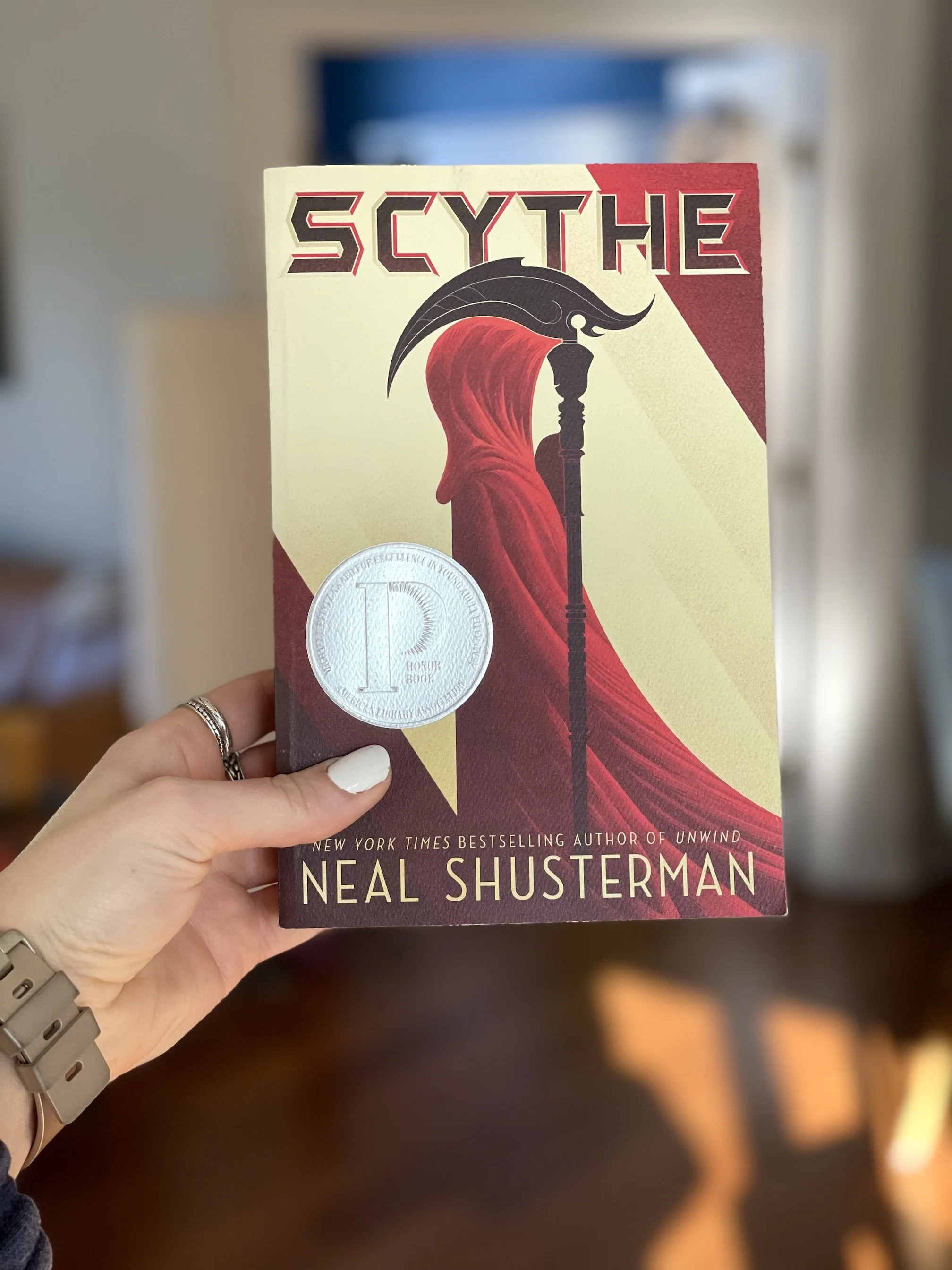 My hand holds a paperback copy of Scythe by Neal Shusterman.