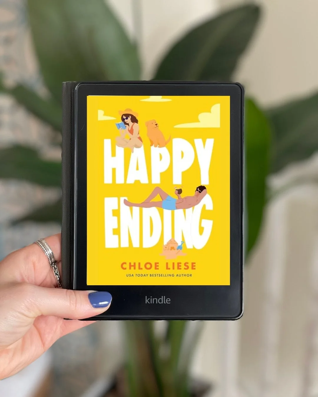 My hand holds a Kindle with the book Happy Ending by Chloe Liese on the screen. The cover is mostly yellow with two people on a beach reading on it.