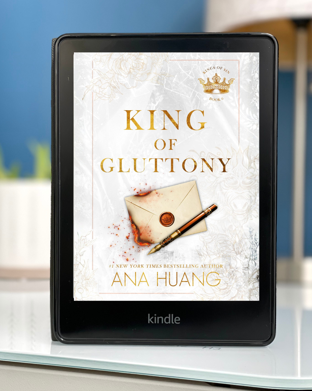 A Kindle rests on a white desk with the cover of King of Gluttony by Ana Huang on the screen. The cover is mostly white with a burning envelope in the middle.