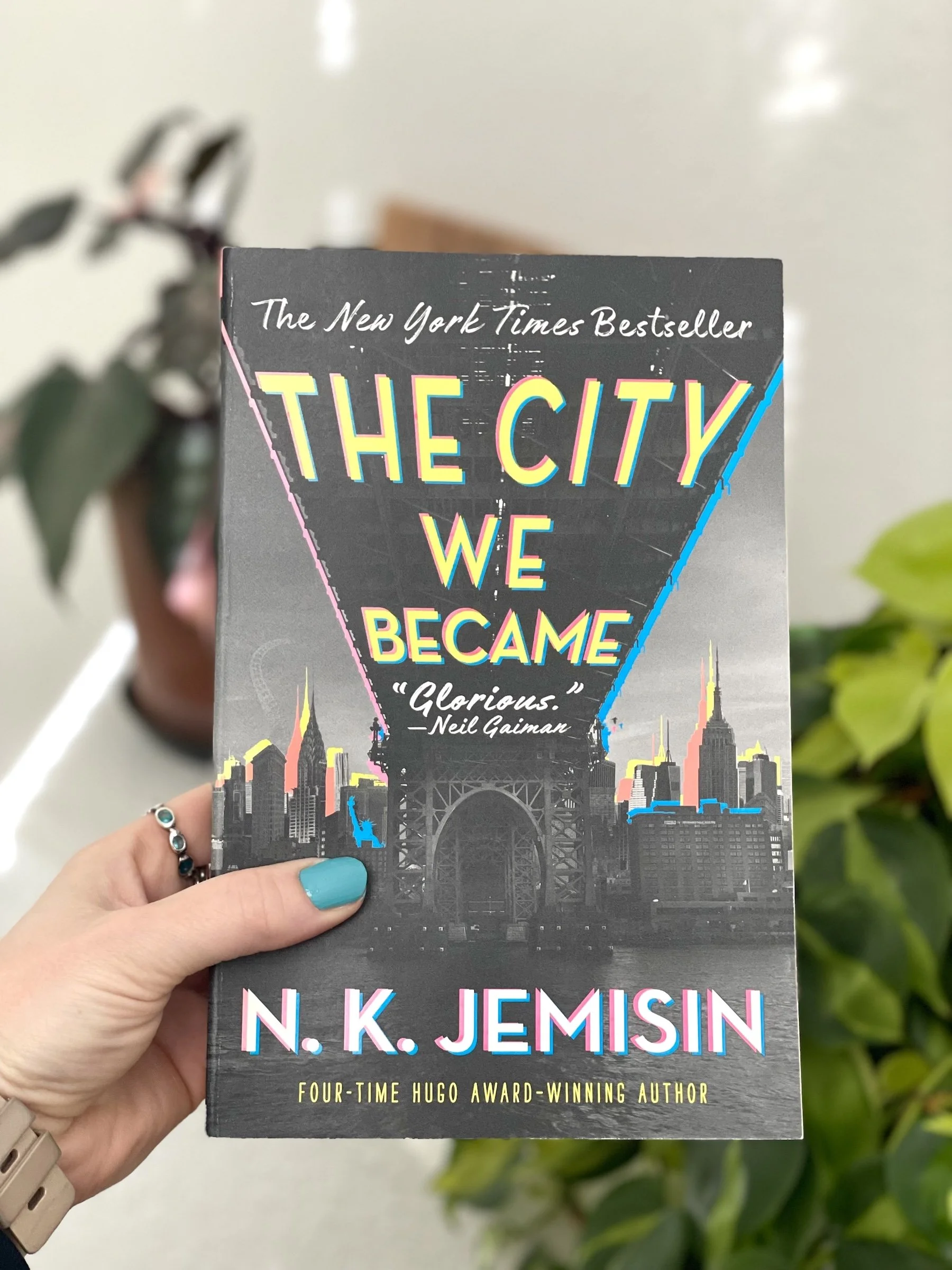 My hand holds a paper back copy of The City We Became by N.K. Jemisin. The cover shows the underside of a bridge in New York City.