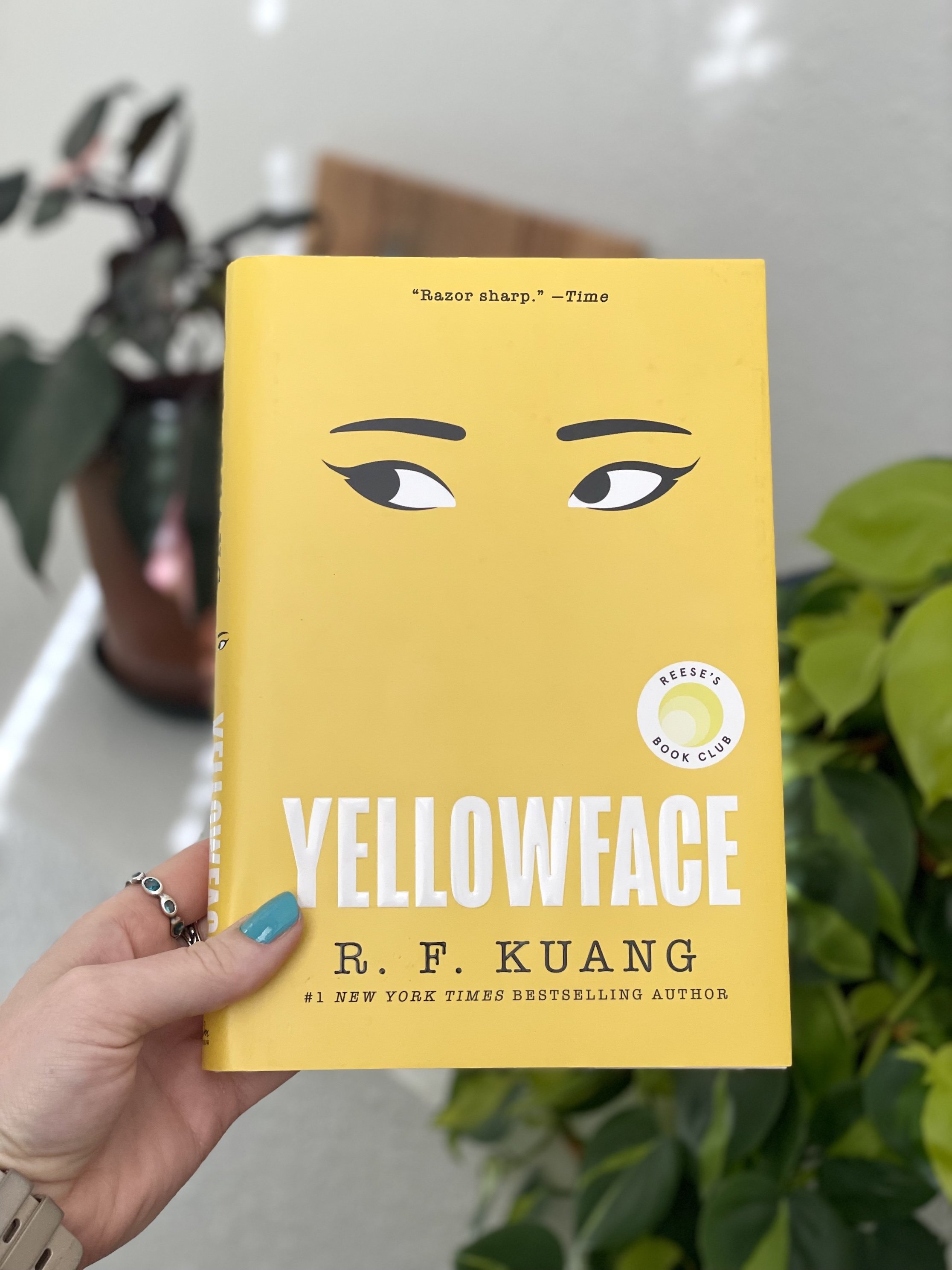 My hand holds a hardcover copy of Yellowface by R.F. Kuang. The cover is mostly yellow with two eyes looking off to the side in the middle.