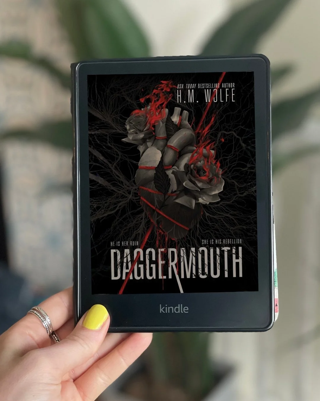 My hand holds a Kindle with the cover of Daggermouth by H.M. Wolfe on its screen. The cover has a gray rose with blood on it.