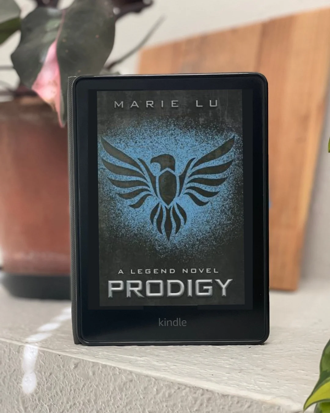 A Kindle with the cover of Prodigy by Marie Lu on the cover. The cover is mostly black with a blue spray painted symbol in the middle.
