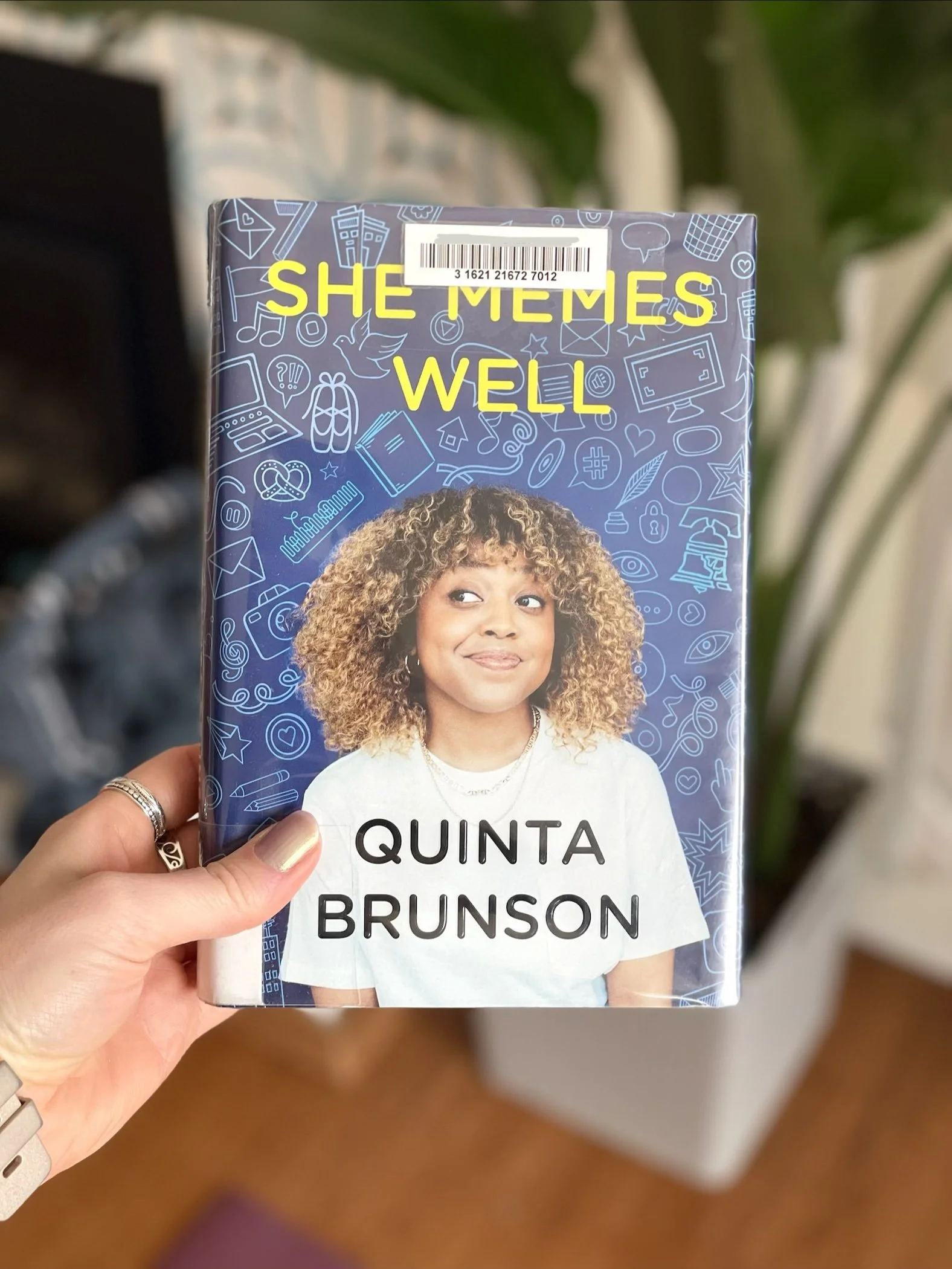 My hand holds a hardcover copy of She Memes Well by Quinta Brunson. The cover is mostly blue with a photo of Quinta in the middle.