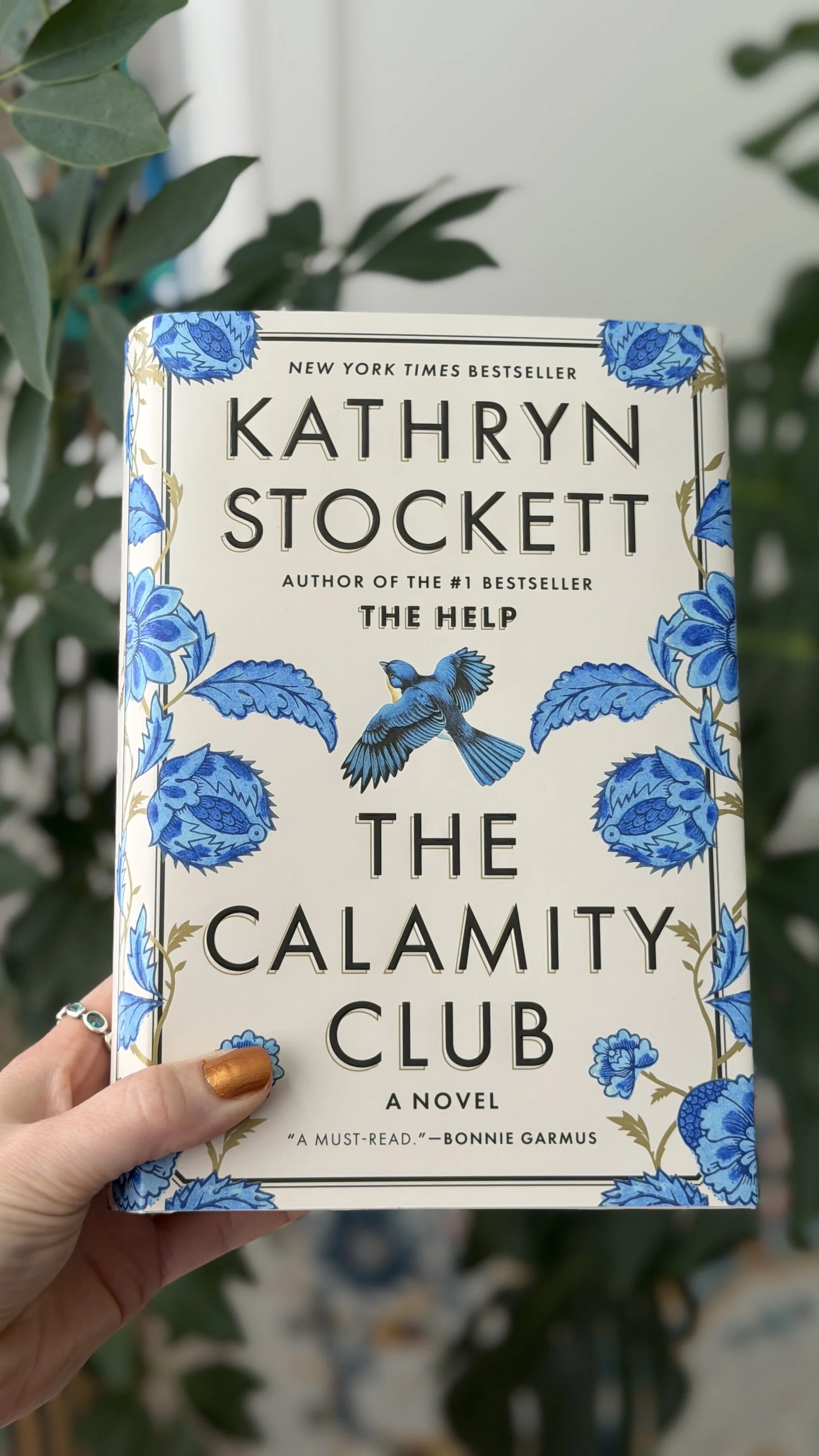 My hand holds a hardcover copy of The Calamity Club by Kathryn Stockett. The cover is mostly cream with blue flowers and a blue bird in the center.