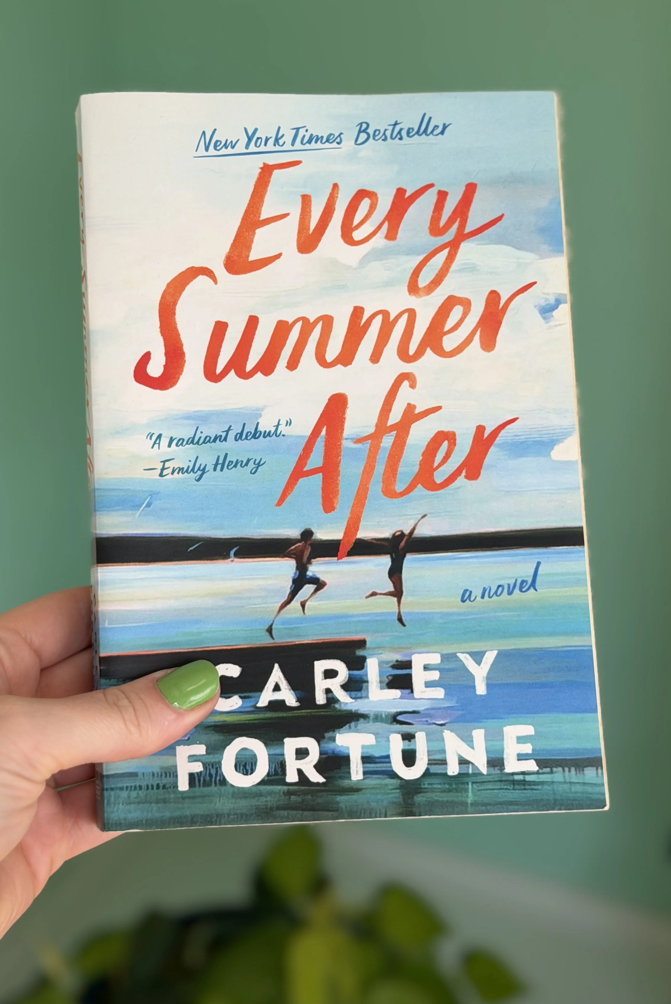 My hand holds a paperback copy of Every Summer After by Carley Fortune. The cover shows two teens jumping off a dock into a lake.