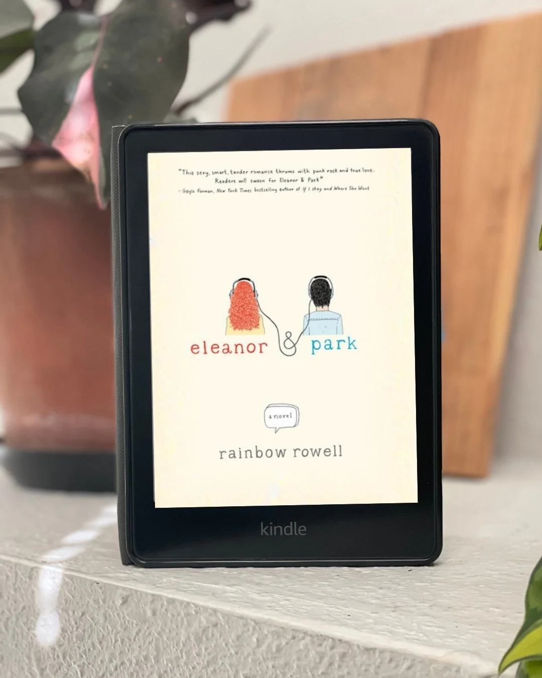 A Kindle with the cover of Eleanor & Park by Rainbow Rowell on the screen. Which shows two teens listening to music.
