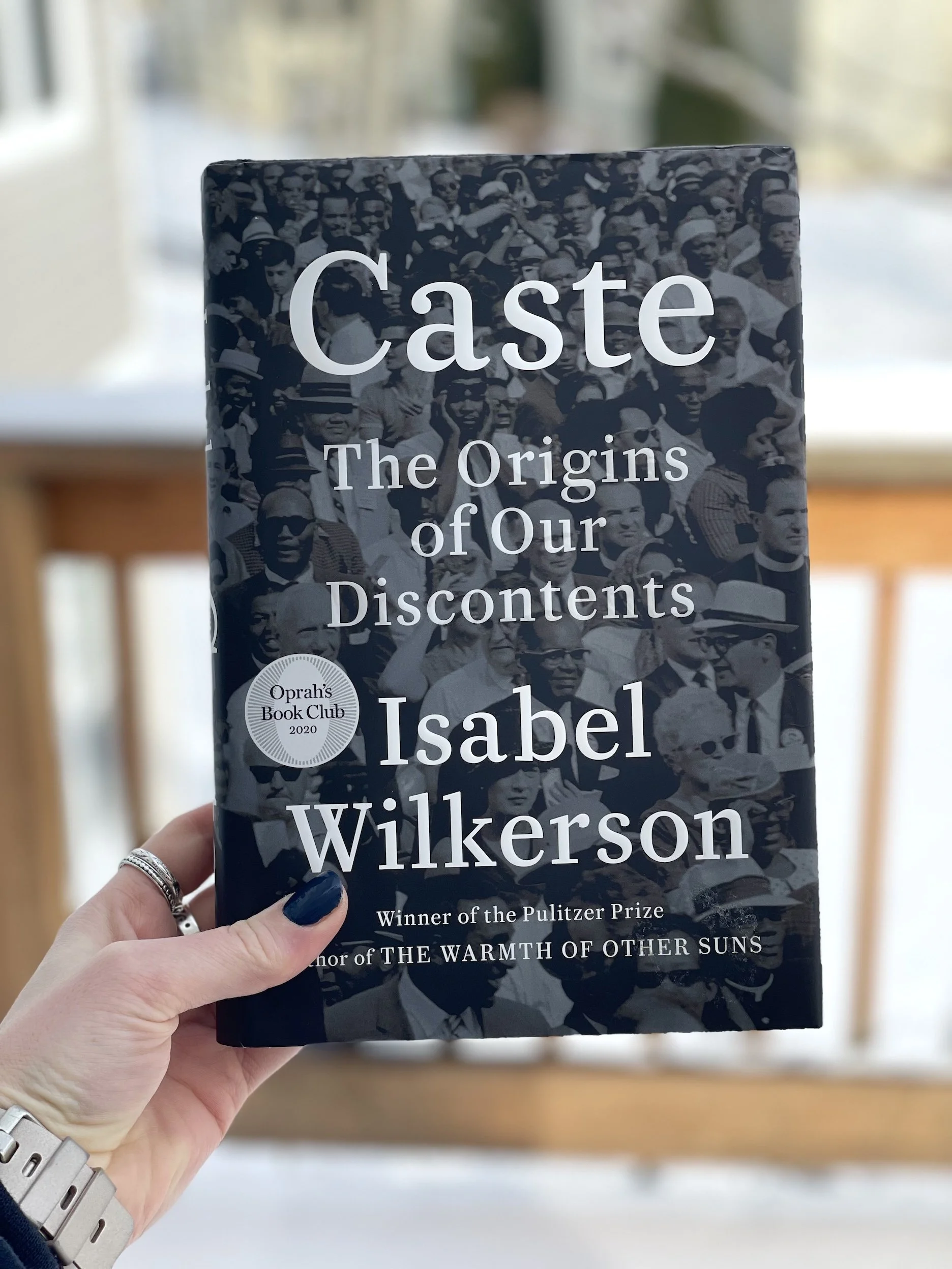 My hand holds a hardcover copy of Caste by Isabel Wilkerson. The cover is mostly black and white.