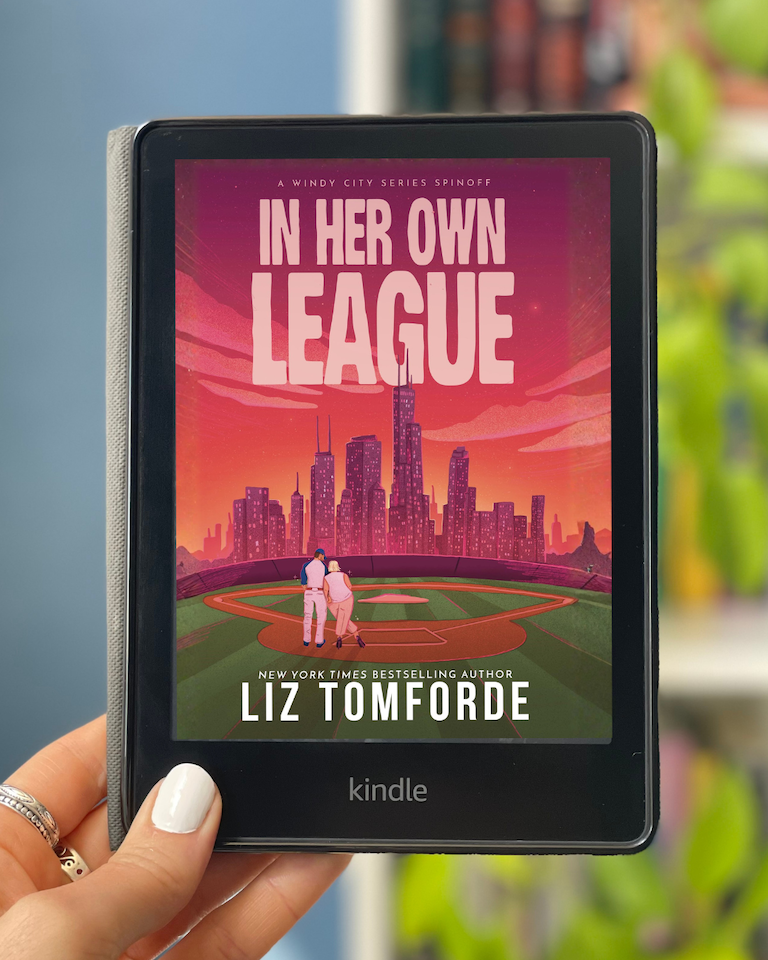 My hand holds a Kindle with the cover of In Her Own League by Liz Tomforde on the screen.