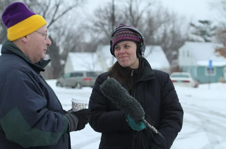 Two people dressed in winter clothing, one holding a coffee cup, standing outside in a snowy neighborhood, with a woman wearing headphones and holding a microphone.