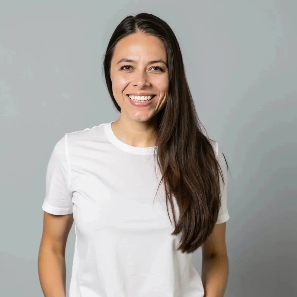 A young woman with long brown hair and a big smile, wearing a white t-shirt, standing against a light gray background.