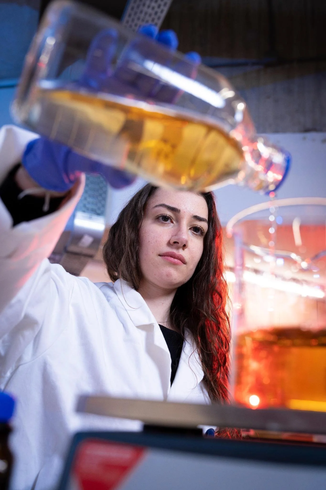 A young woman in a white lab coat and gloves holding a beaker of amber-colored liquid in a laboratory setting.
