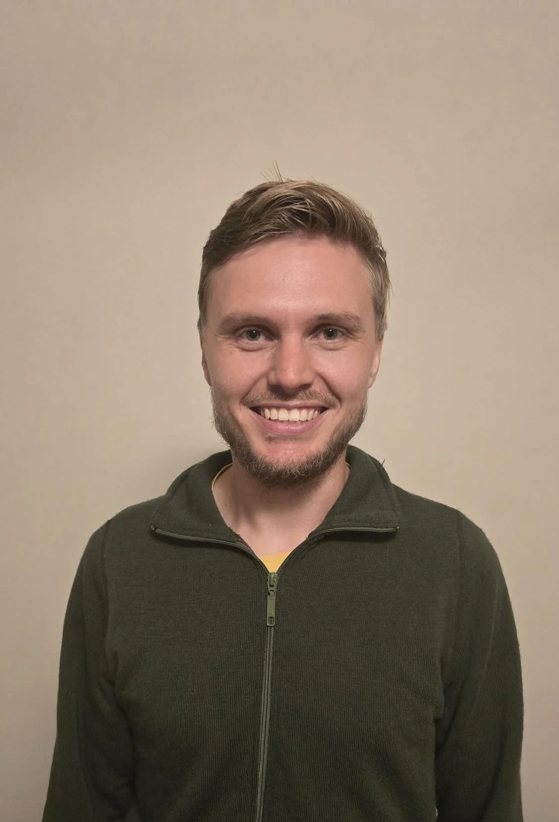 A young man with light skin, short brown hair, and a beard, smiling in front of a plain beige wall.