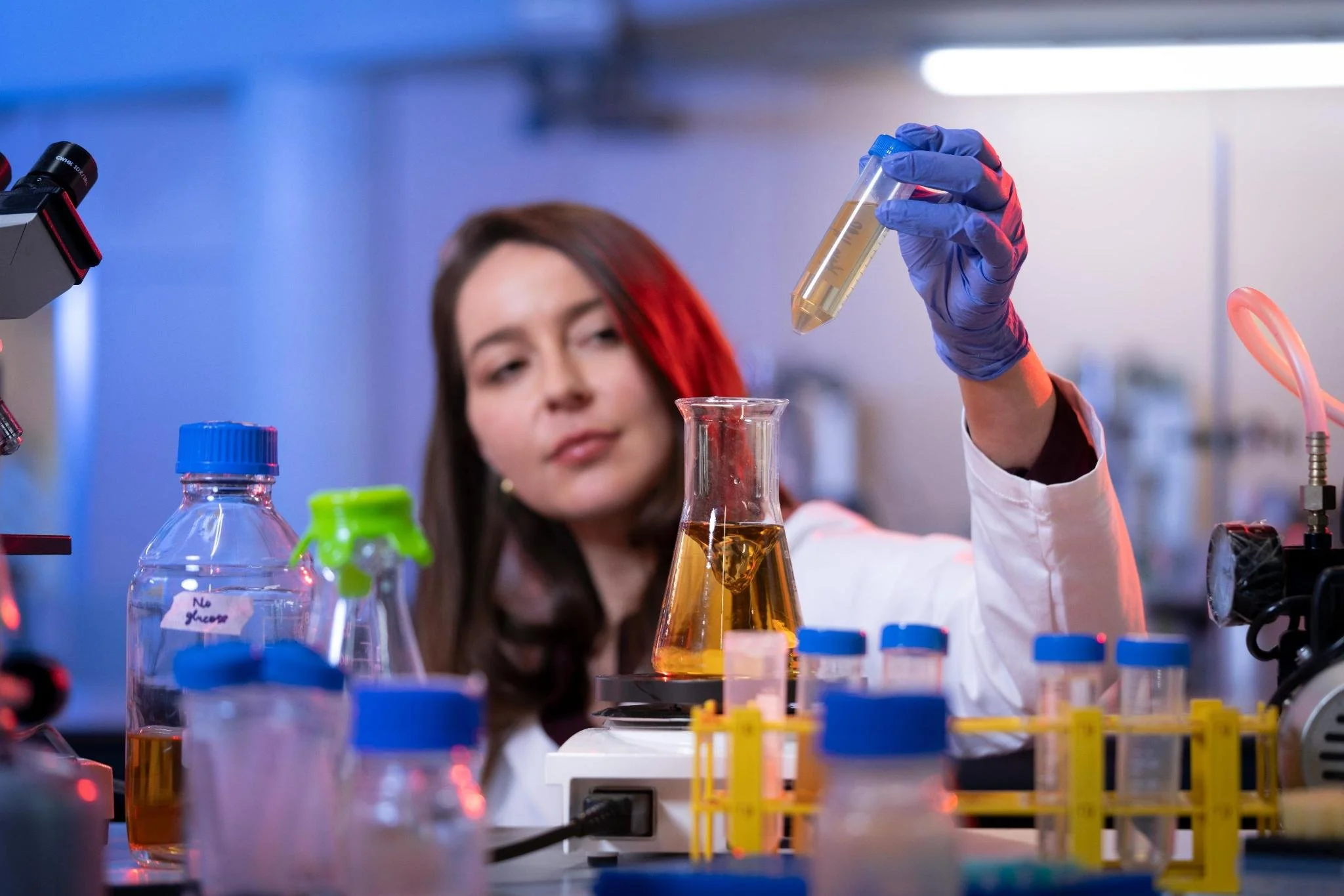 A female scientist wearing gloves holds a test tube with a yellow liquid in a laboratory filled with various lab equipment.