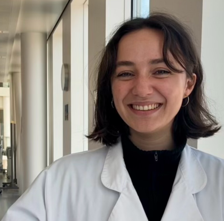 A young woman with dark, shoulder-length hair and silver hoop earrings smiling in a bright, modern building with large windows and light-colored walls, wearing a white lab coat over a black top.