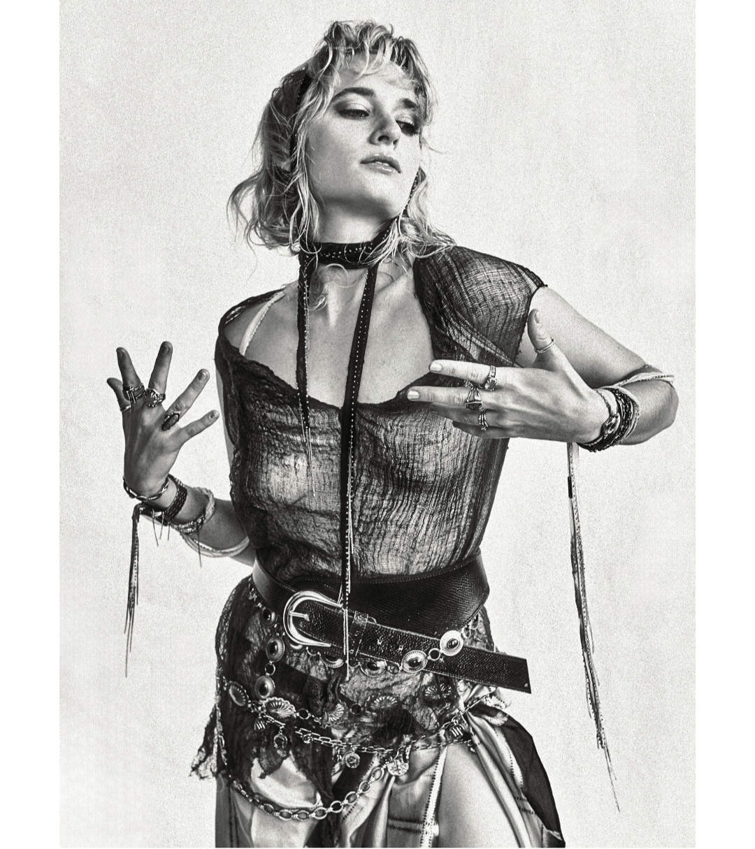 Lorian Gish by Emmanuel Rosario. Blonde figure draped in beaded ribbons, wrapped with belts and chains, gesturing and looking down towards the website text