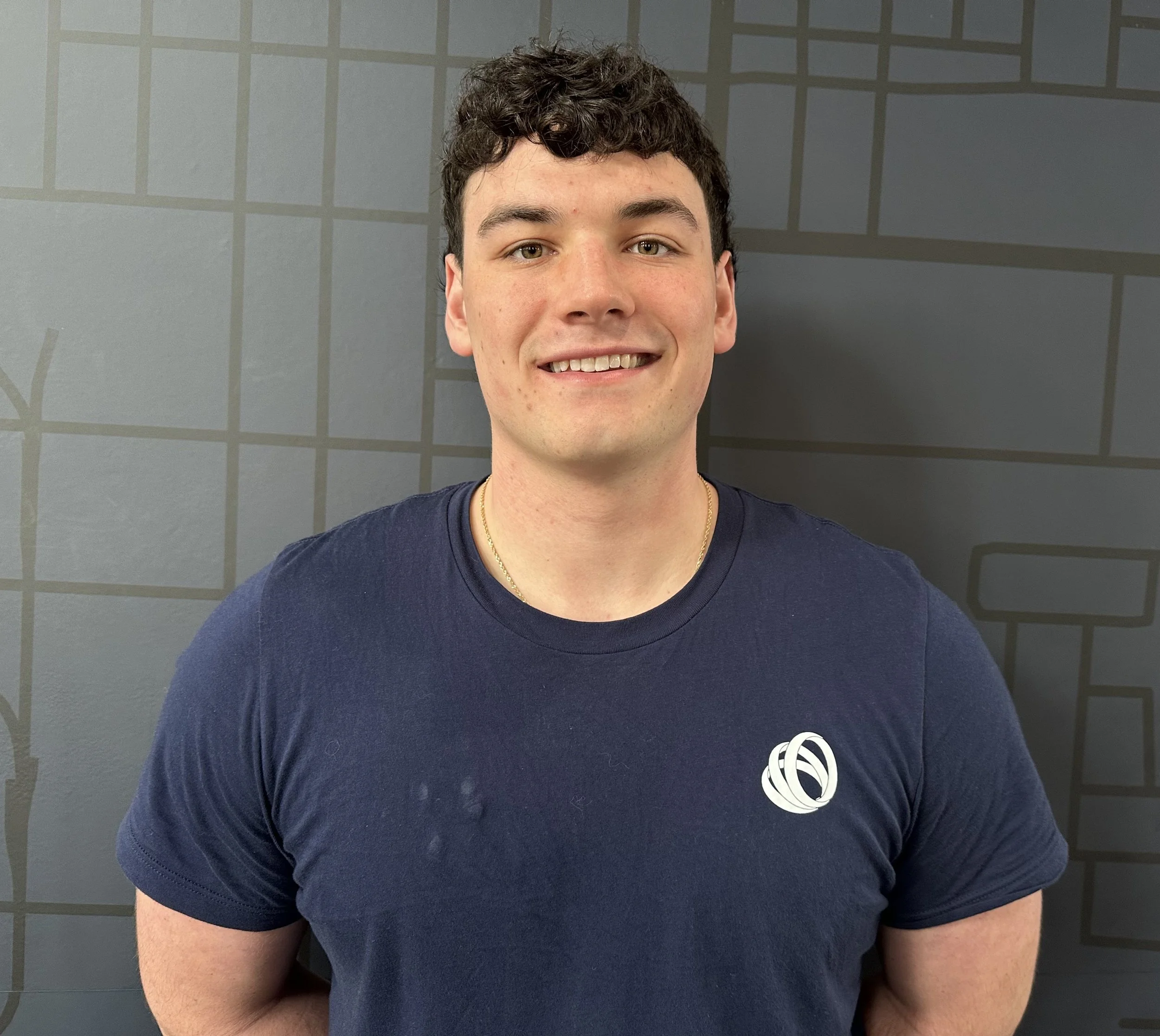 A young man with short dark curly hair and a light complexion, smiling, wearing a navy blue t-shirt with a white circular logo on the chest, standing against a dark gray wall with a subtle geometric pattern.