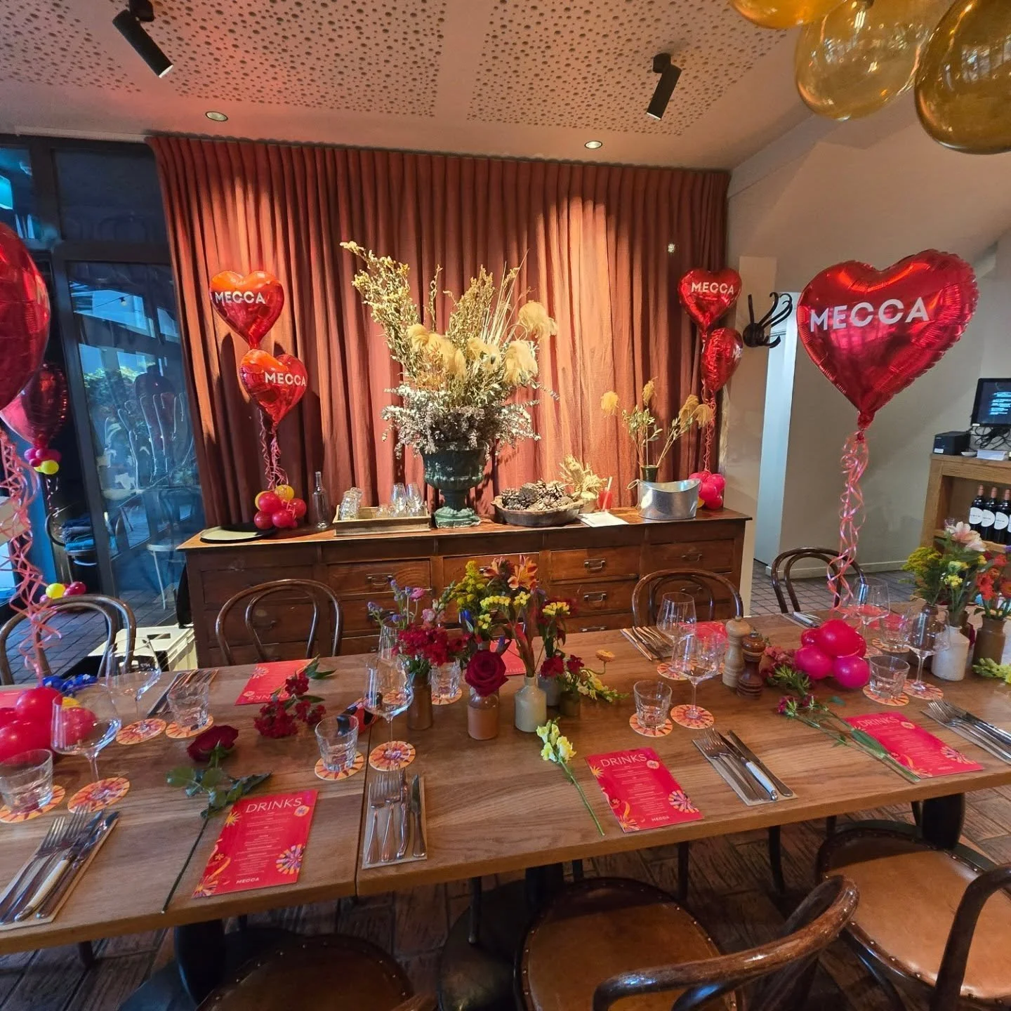 A decorated indoor celebration table with red and pink balloons with the word 'Mecca,' floral arrangements, and a wooden sideboard in the background, set for a festive occasion.
