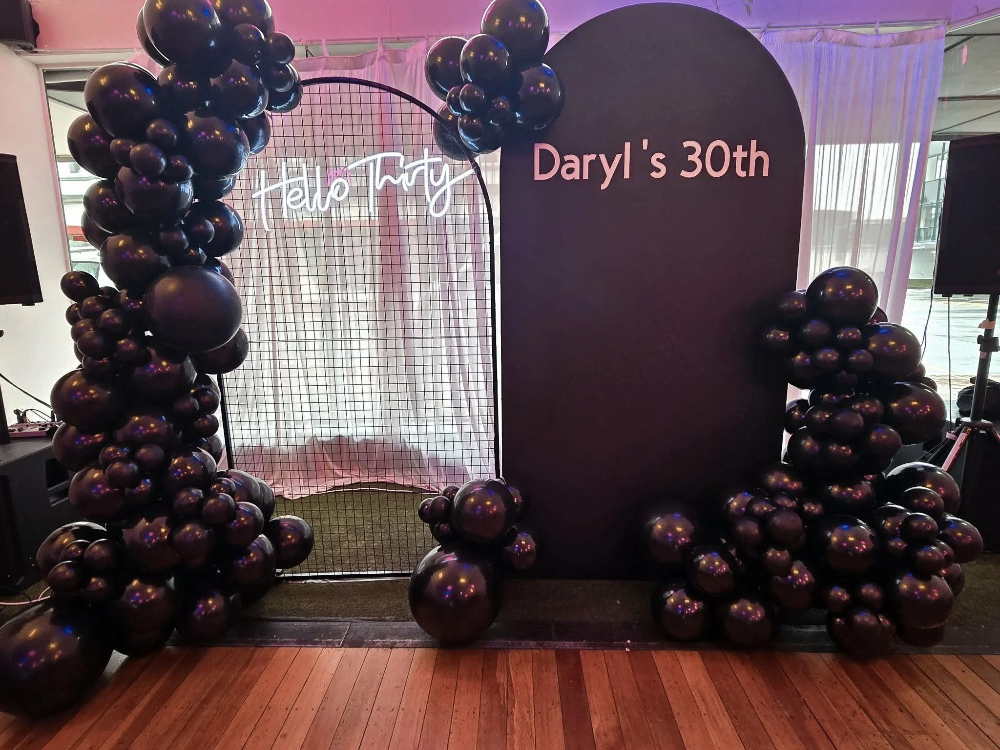 Decorative birthday celebration setup with black balloons clustered around a black backdrop and a wire grid with pink curtains, celebrating Daryl's 30th birthday.