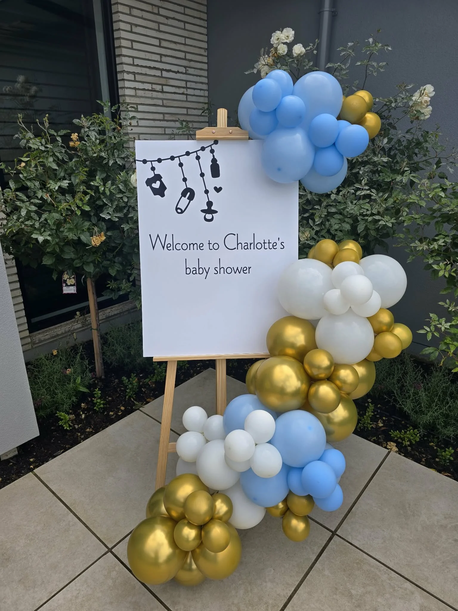 Decorative baby shower sign with black illustrations and text, surrounded by blue, white, and gold balloons, set outdoors near bushes and a building.