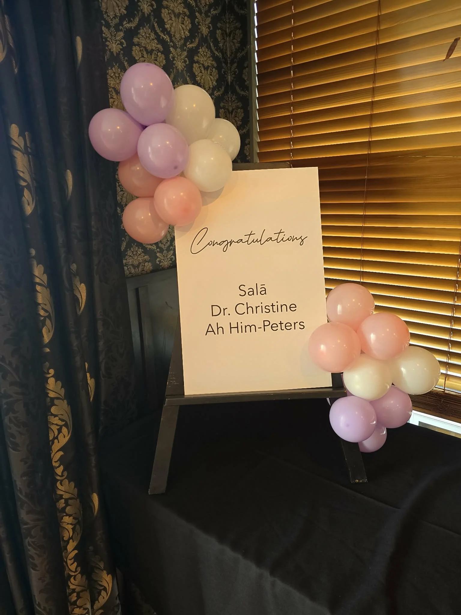 A congratulatory sign with balloons on a table, in a room with dark floral wallpaper and wooden blinds, celebrating Dr. Christine Ah Him-Peters.