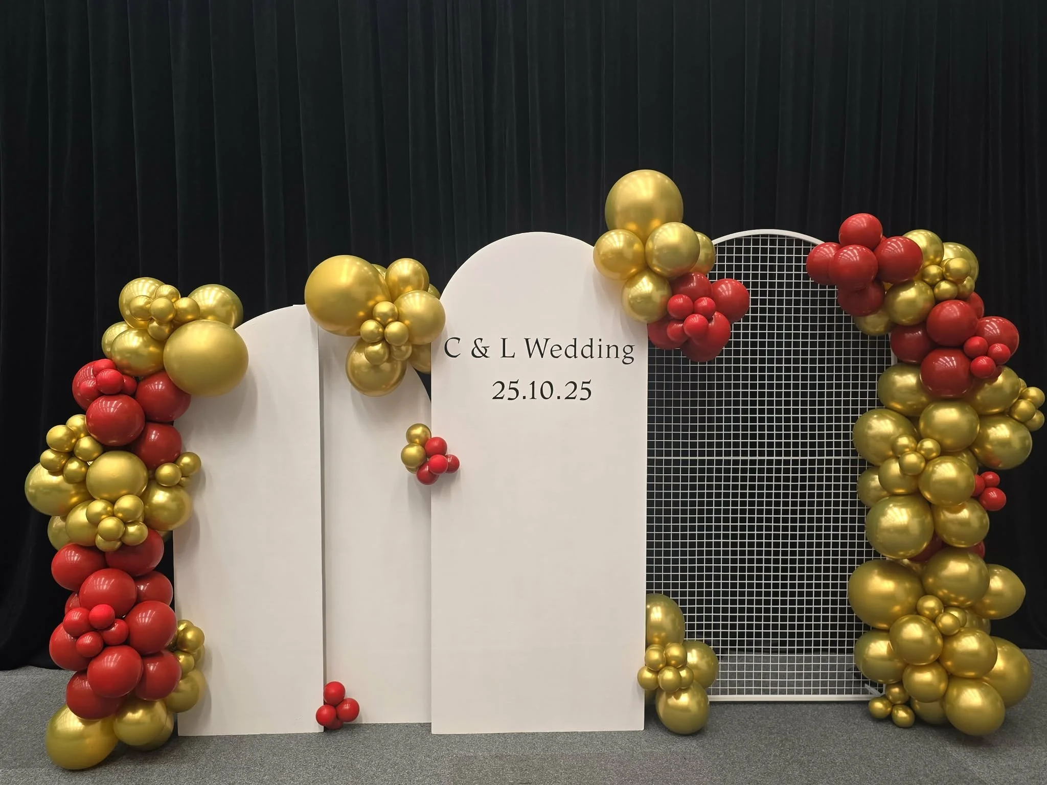 Decorative wedding backdrop with white panels, gold and red balloons, and a sign reading 'C & L Wedding 25.10.25'.