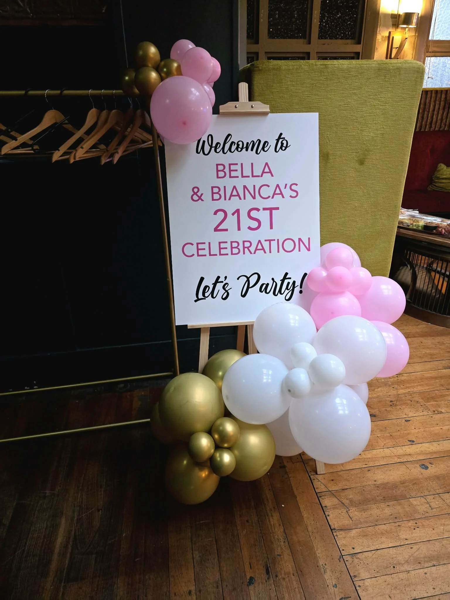 A welcome signboard for Bella & Bianca's 21st celebration party, decorated with pink, white, and gold balloons, placed on a wooden floor near a green chair and a clothing rack.
